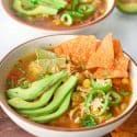 ¾ angle photo of chicken tortilla soup with sliced avocado, jalapeño and tortilla chips on top.