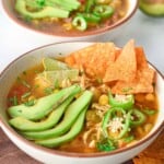 ¾ angle photo of chicken tortilla soup with sliced avocado, jalapeño and tortilla chips on top.
