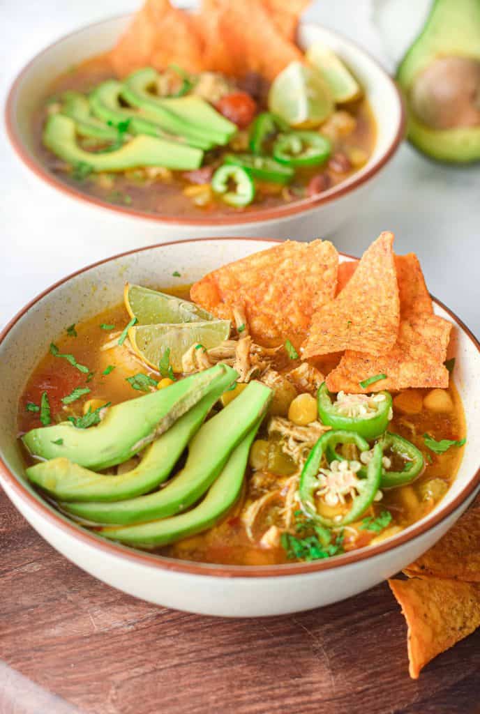 ¾ angle photo of chicken tortilla soup with sliced avocado, jalapeño and tortilla chips on top.