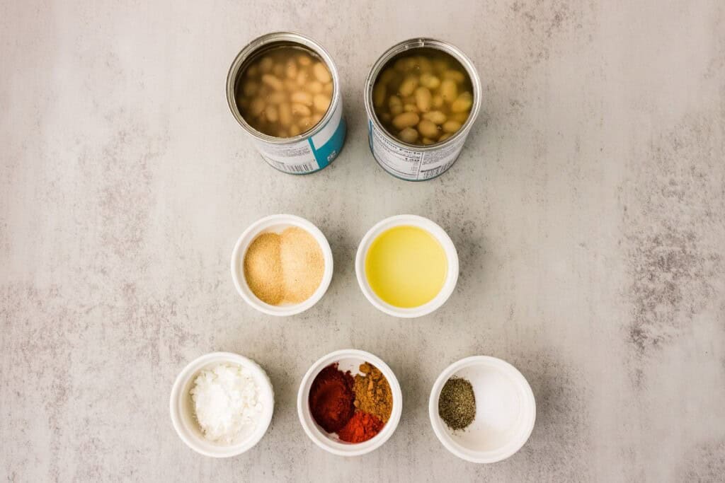 Crispy white beans ingredients flat lay with two cans of great northern beans, olive oil, and pantry spices in small white bowls.