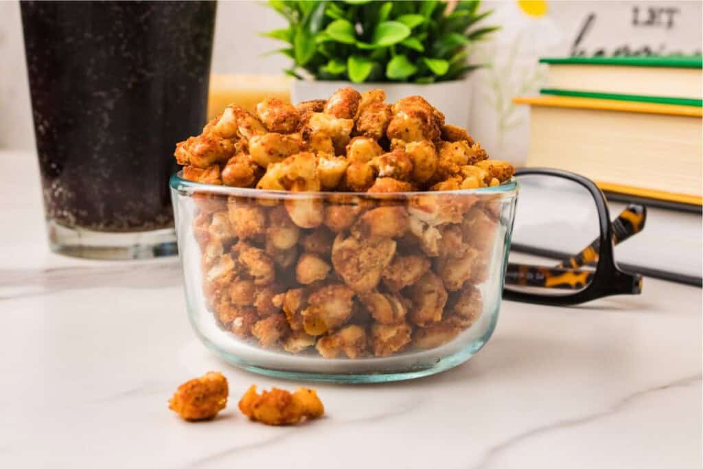 Crispy white beans in a small glass bowl on a marble surface with a cola, plant, and stacked books - a snack alternative to chips.