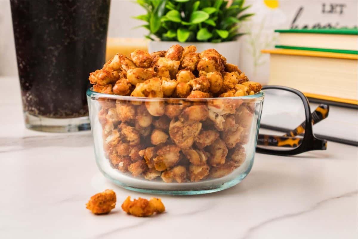 Crispy white beans in a small glass bowl on a marble surface with a cola, plant, and stacked books — a snack alternative to chips.