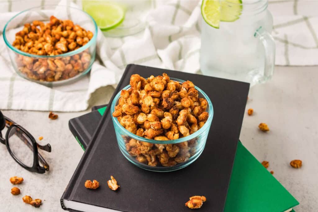 Crispy roasted white beans in a glass bowl on a stack of books with reading glasses and a glass of seltzer - a high-protein snack alternative to chips.