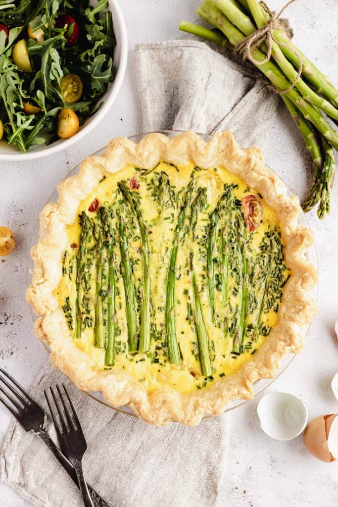Asparagus and tomato quiche on a white table cloth with a salad on the left and asparagus stalks on the right.