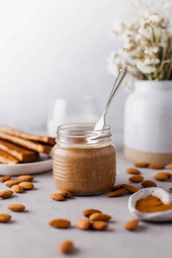 Almond butter in glass Mason Jar with spoon in it. Slices of toast and vase of dried flowers behind it.