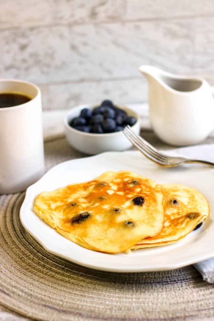 Blueberry pancakes on a white plate with a bowl of blueberries behind it and a creamer pitcher.