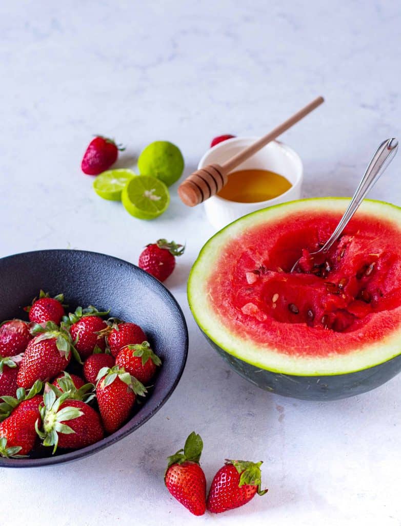 Half of a watermelon with a spoon in it, bowl of strawberries, honey and lime cut in half.