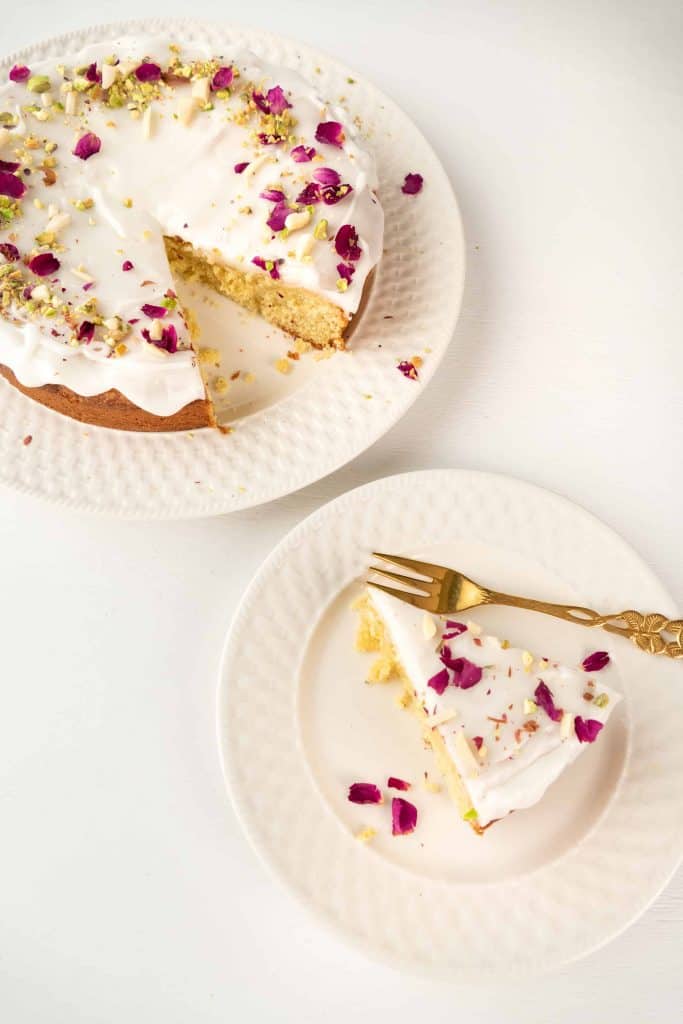Overhead shot of Persian Love Cake with a slice cut out of it and a piece on a white plate on the side with a gold fork on the plate.