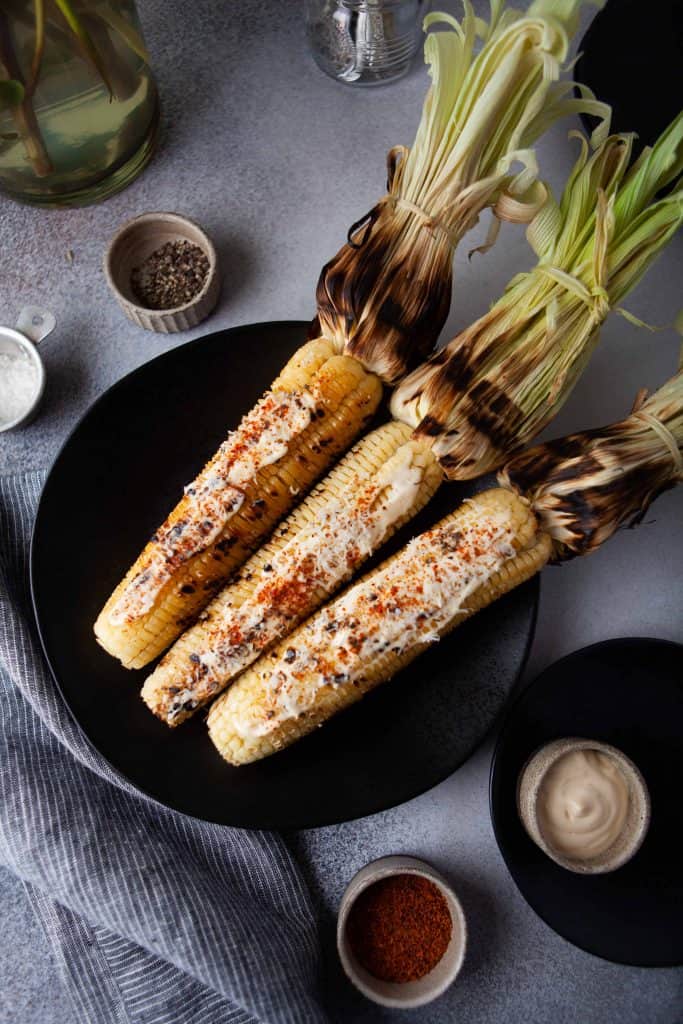 Grilled corn on the cob on a black plate with spices around it.