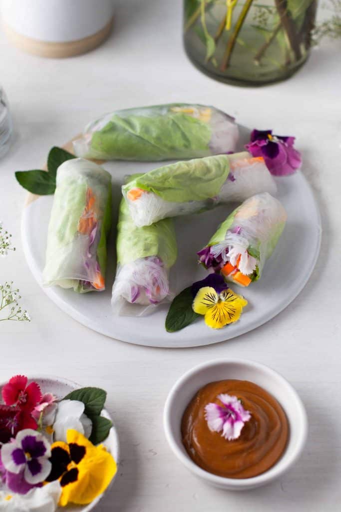 Plate of spring rolls with bowl of edible flowers and bowl of peanut sauce beside it.