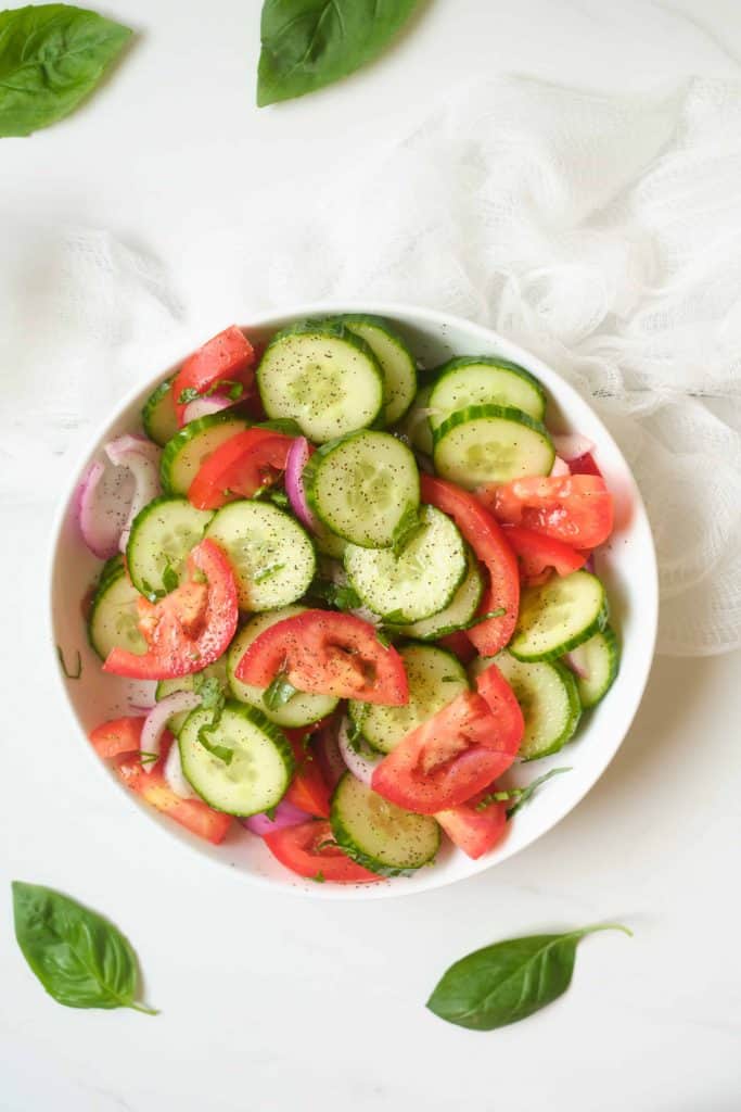 Large white bowl of sliced cucumbers, tomato, red onion and basil.
