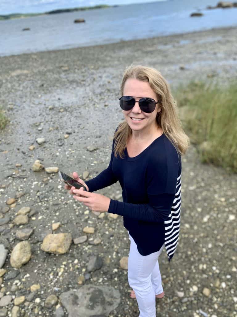 Pretty blonde lady wearing sunglasses on the beach holding cell phone.