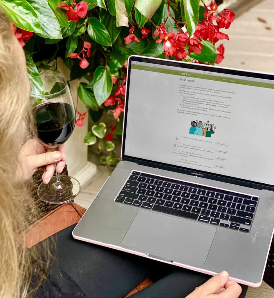 Blonde lady holding a glass of red wine while looking at her laptop.