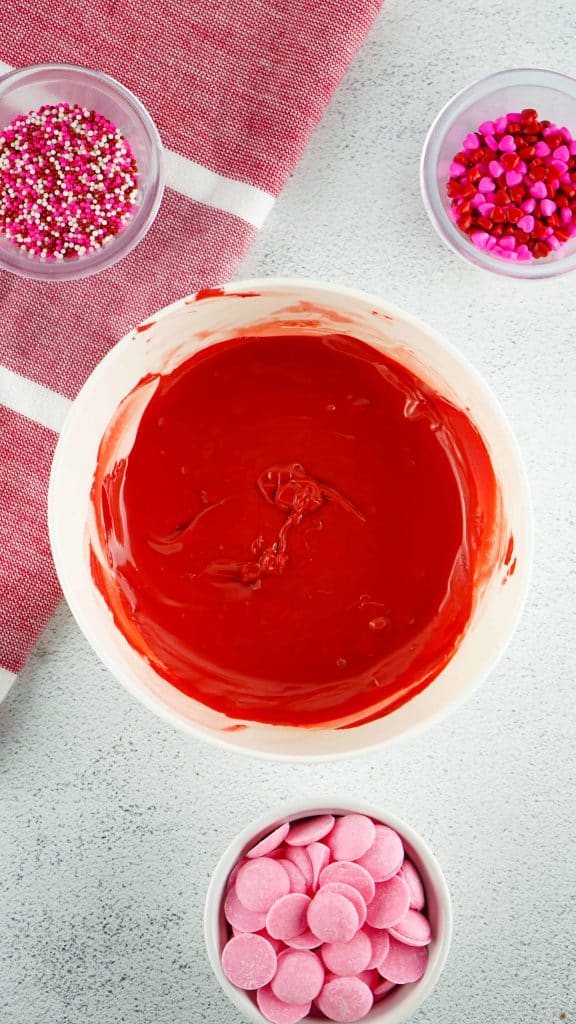 Melted red candy melts in a white bowl with bowls of cookie decorations around it.