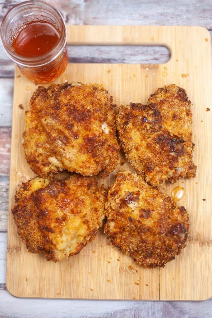 Cooked, breaded chicken thighs on a cutting board with a jar of honey glaze one the side.