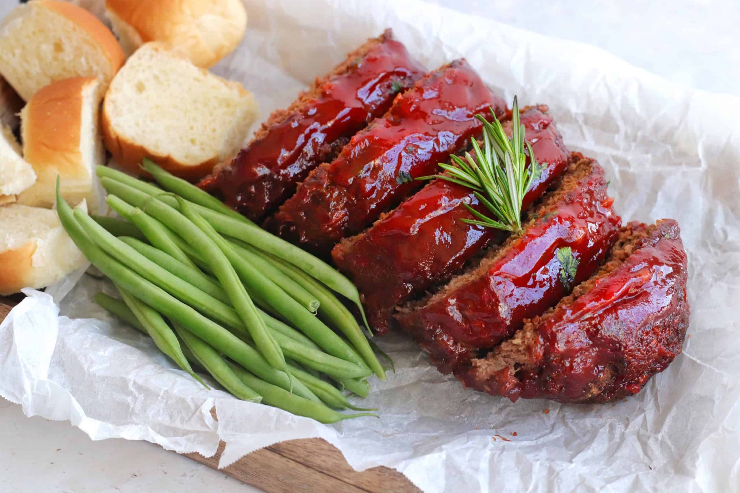 Air fryer meatloaf with glaze on a parchment-lined platter with rosemary sprigs on top, green beans and rolls.