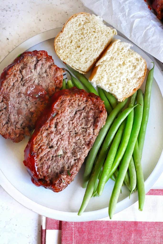 Overhead view of meatloaf with green beans and roll cut in half on a white plate.