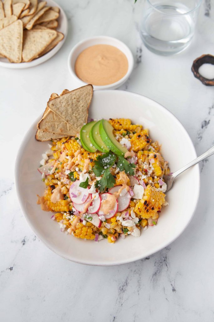 Corn salad with tortilla chips, avocado slices and radish slices on top of it.