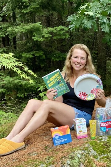 Lady sitting outside on a rock holding a book and paper plates with other products in front of her.