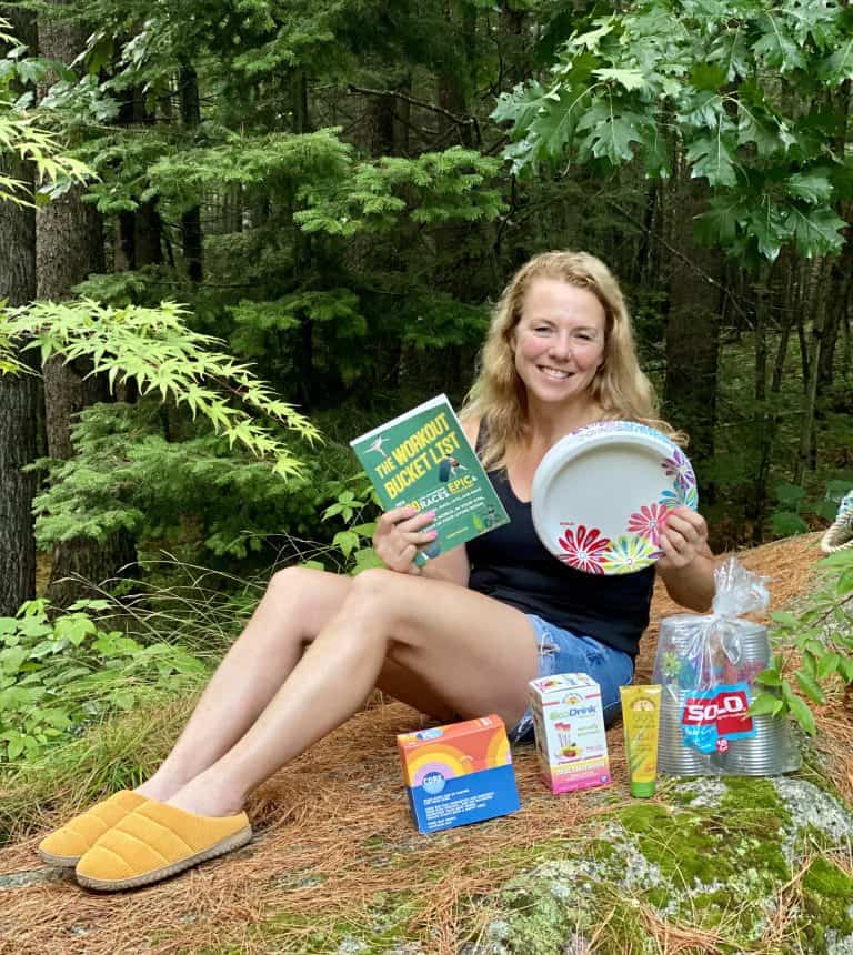 Lady sitting outside on a rock holding a book and paper plates with other products in front of her.