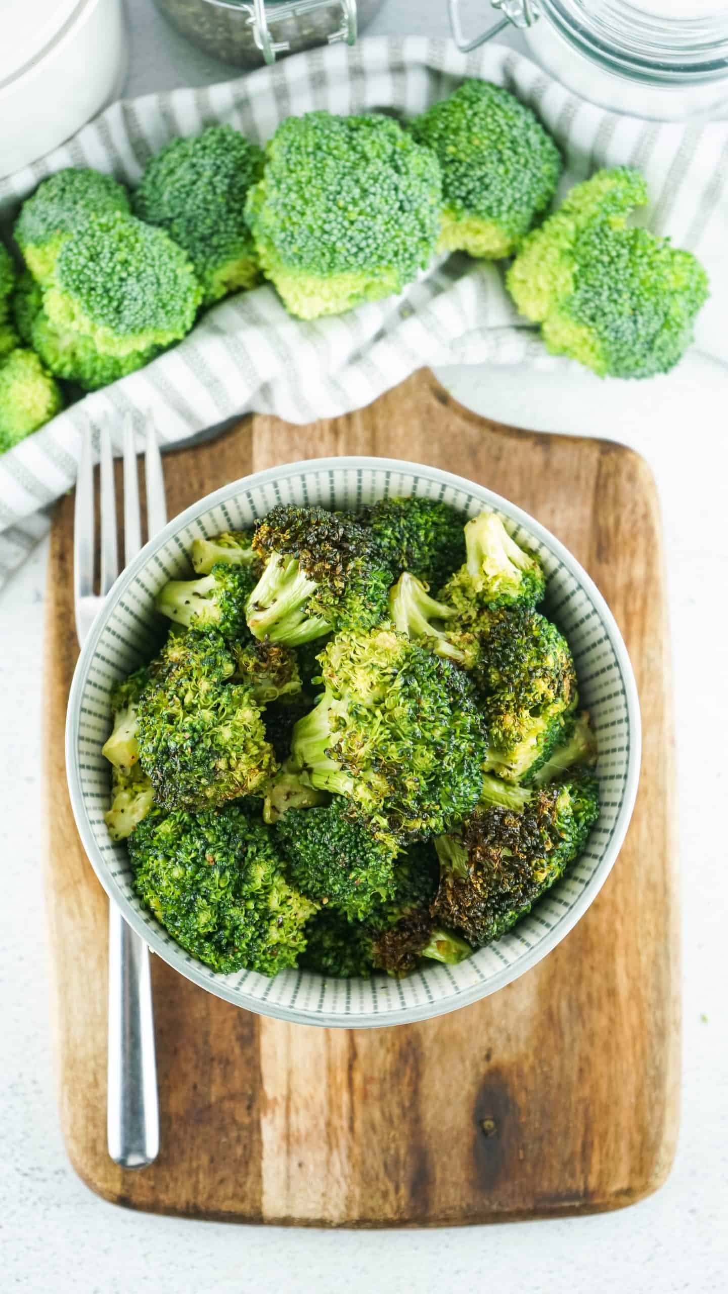 Overhead view of cooked broccoli in a bowl on top of cutting board. Fresh broccoli at top of the photo.