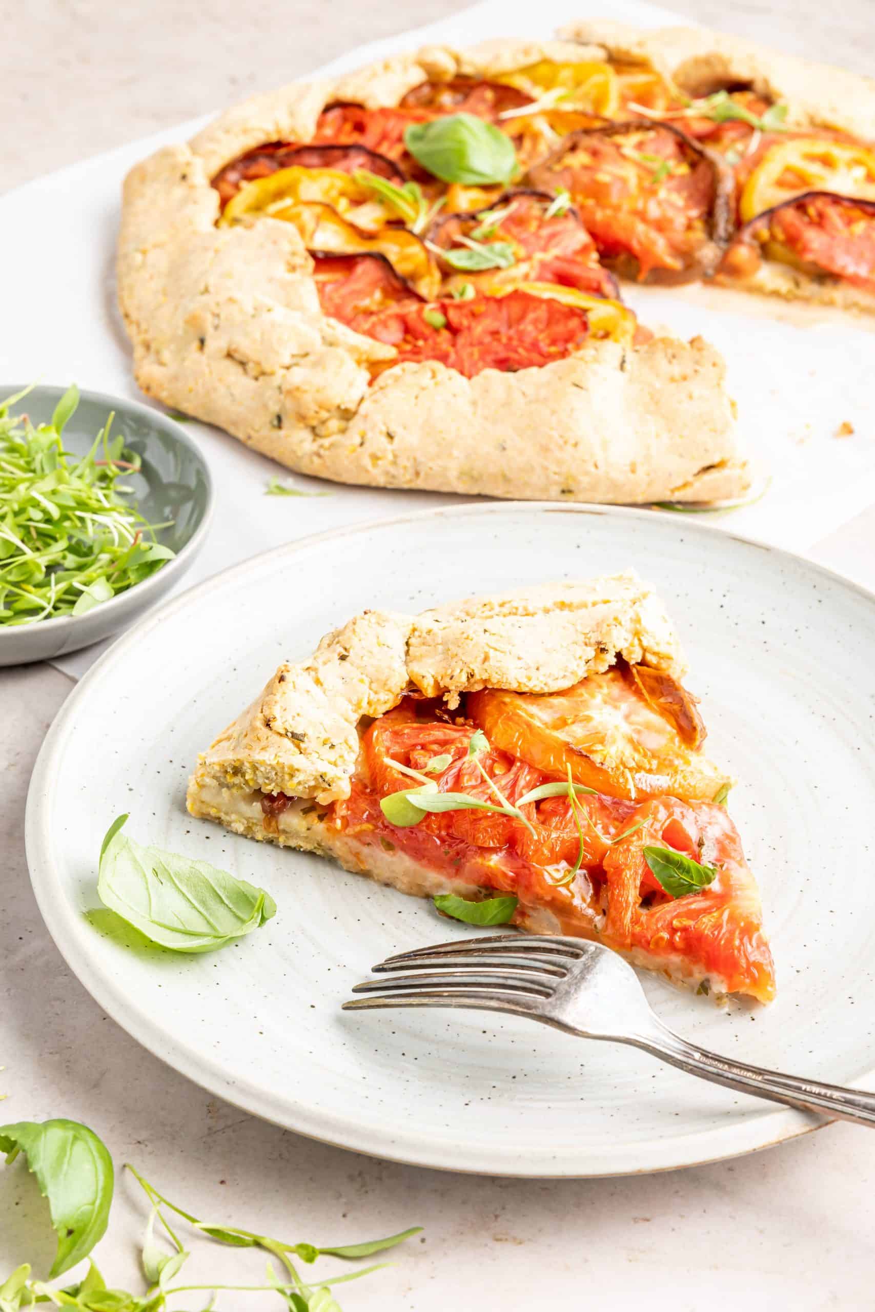 Slice of tomato galette on plate with heirloom tomato galette behind it and a bowl of fresh green herbs.