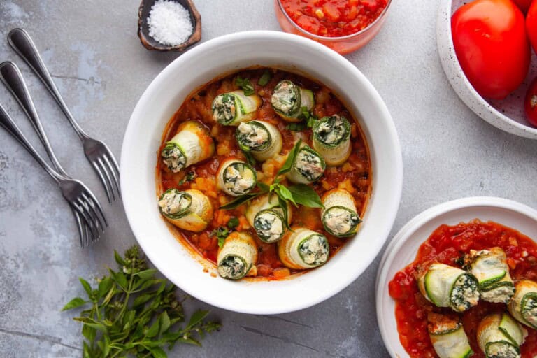 Overhead view of a serving of zucchini lasagna rolls in a white dish with silverware next to it.