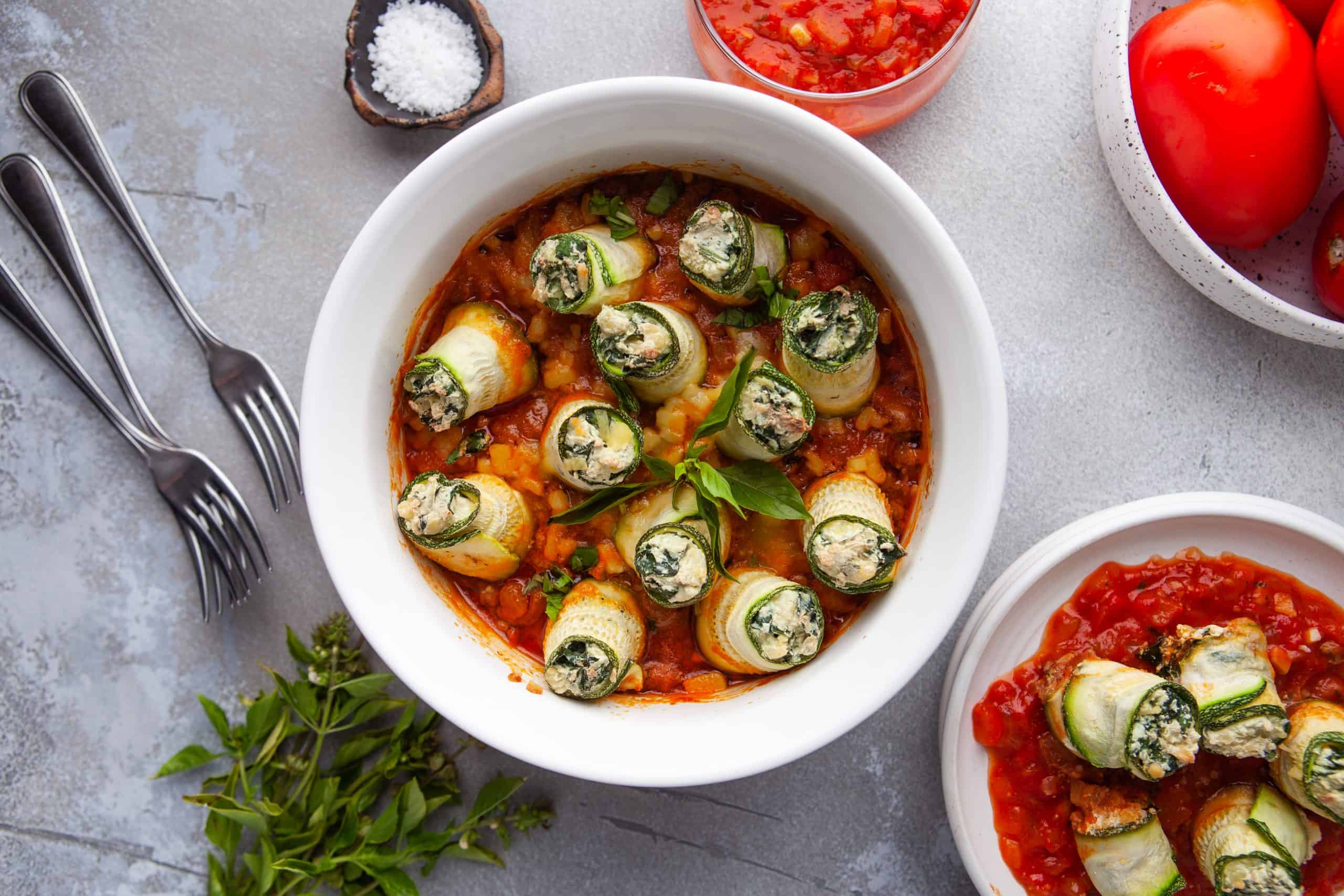 Overhead view of a serving of zucchini lasagna rolls in a white dish with silverware next to it.