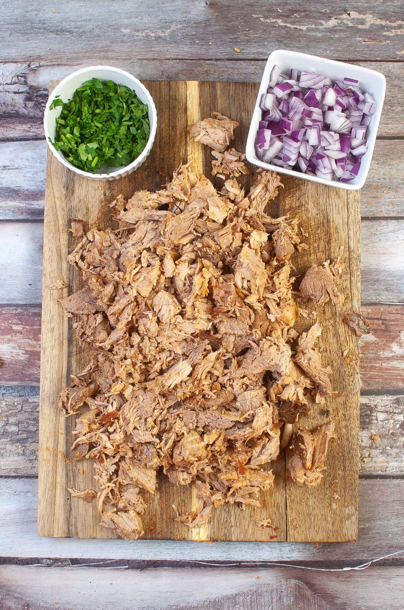 Shredded pork on cutting board with cilantro and diced red onions.