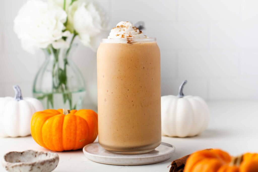 Smoothie with pumpkin spice and whipped cream on a white counter with mini pumpkins behind it.