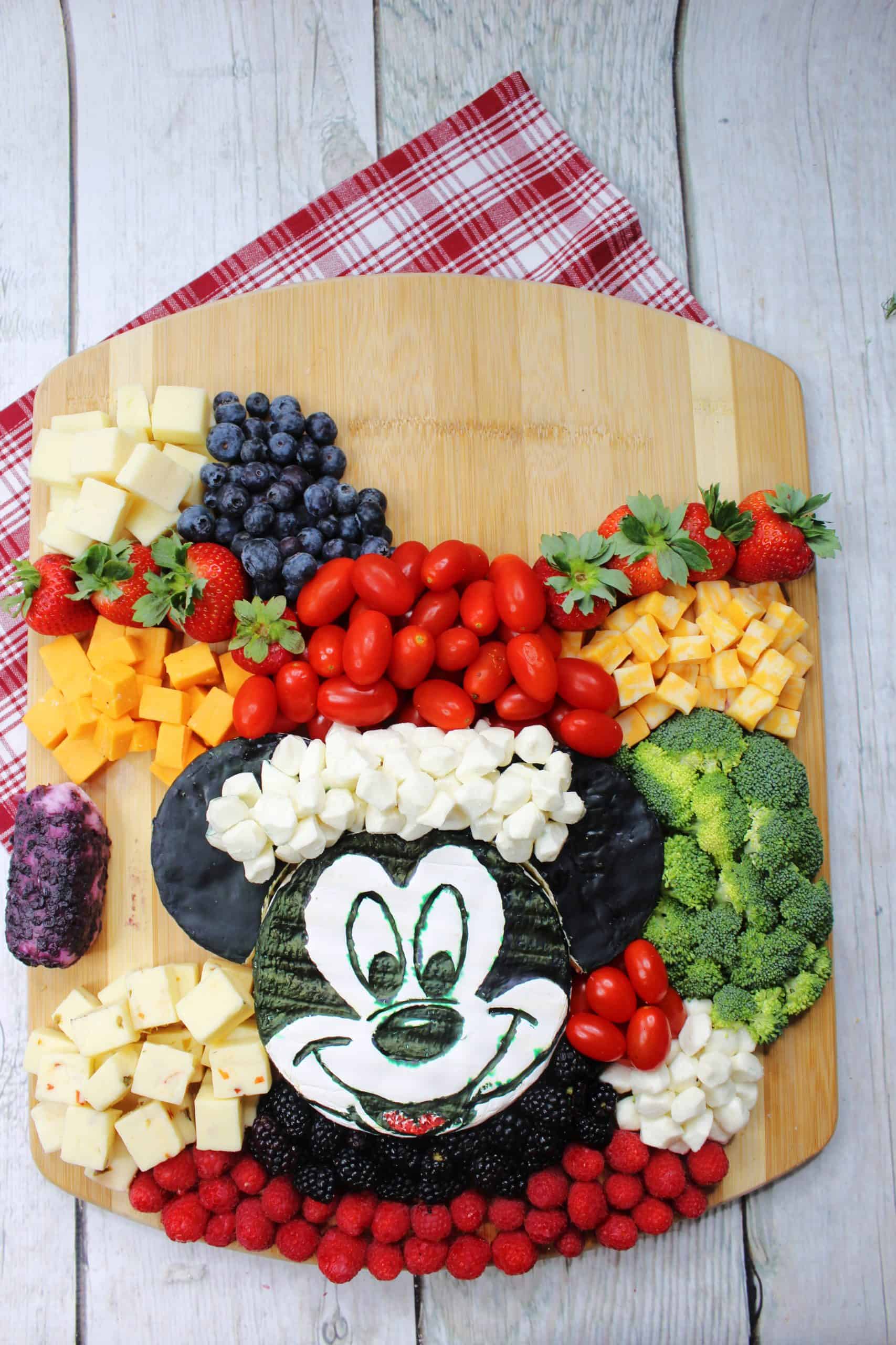 Adding fruit and veggies around Santa Mickey Brie on wooden board.