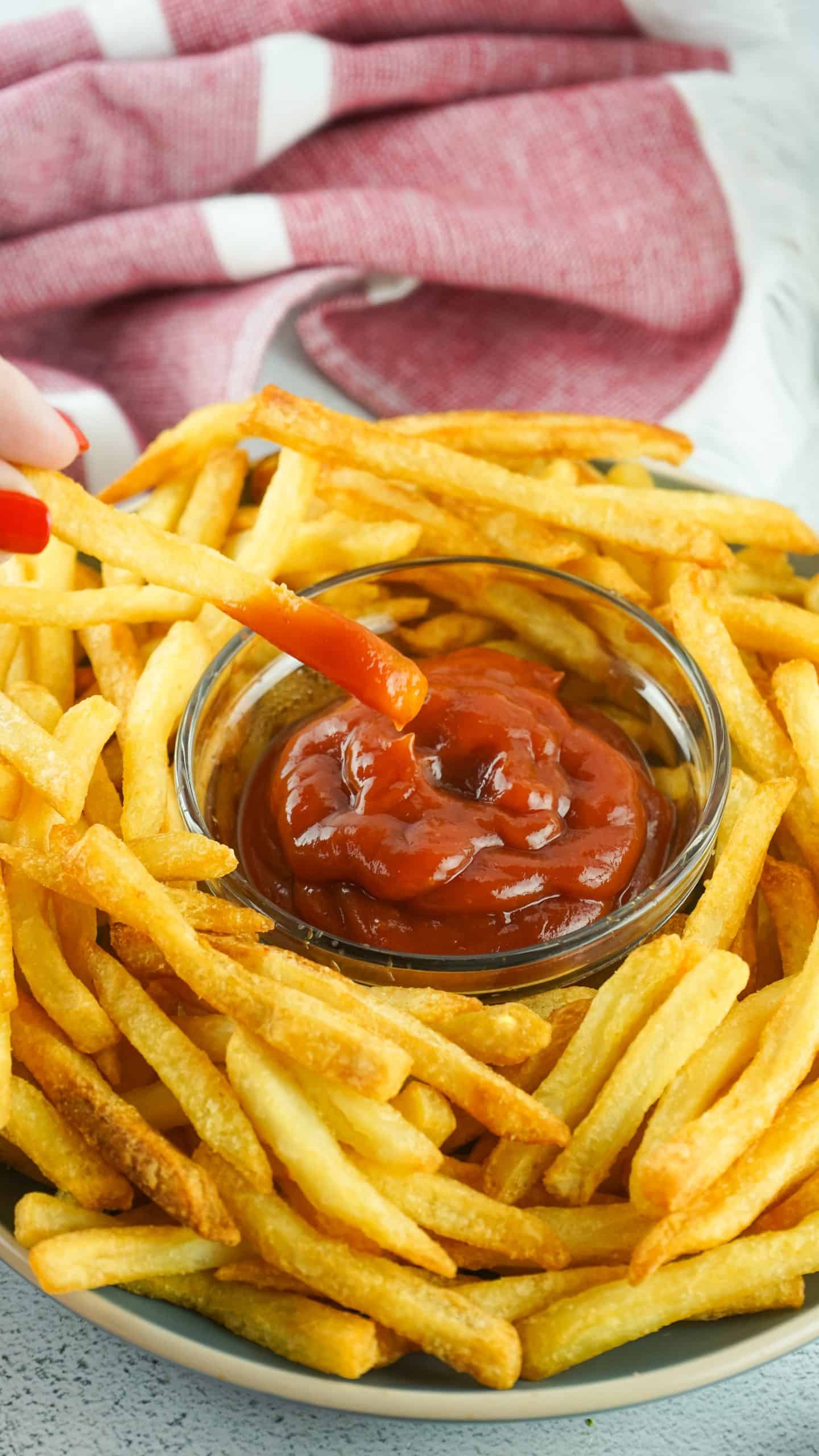 Closeup of plate of French fries and glimpse of hand while dipping. one fry into bowl of ketchup in center of plate.