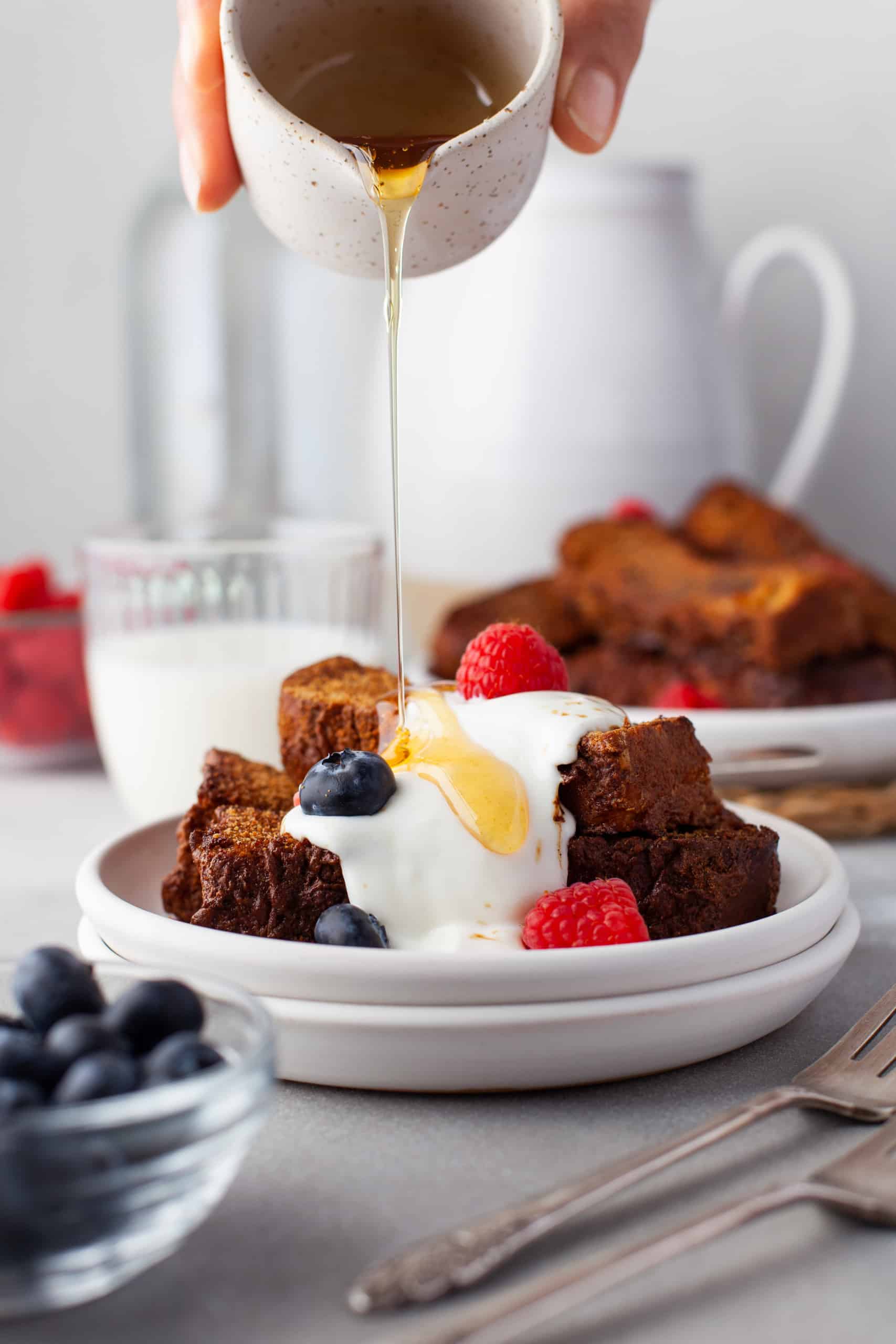 Pouring maple syrup onto French toast sticks with cream and fresh berries.
