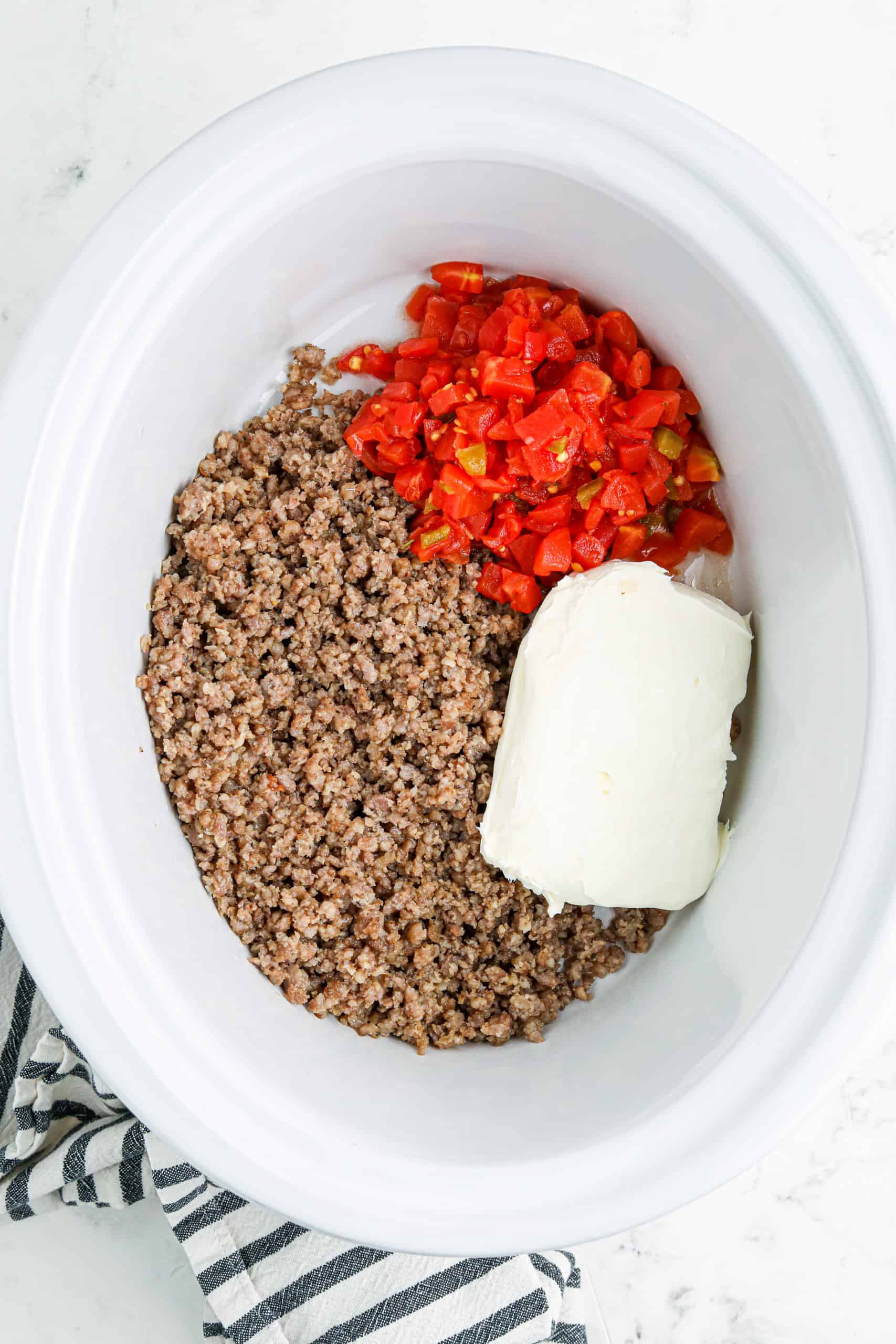 Overhead view of cream cheese, browned sausage and Rotel in a white crockpot.