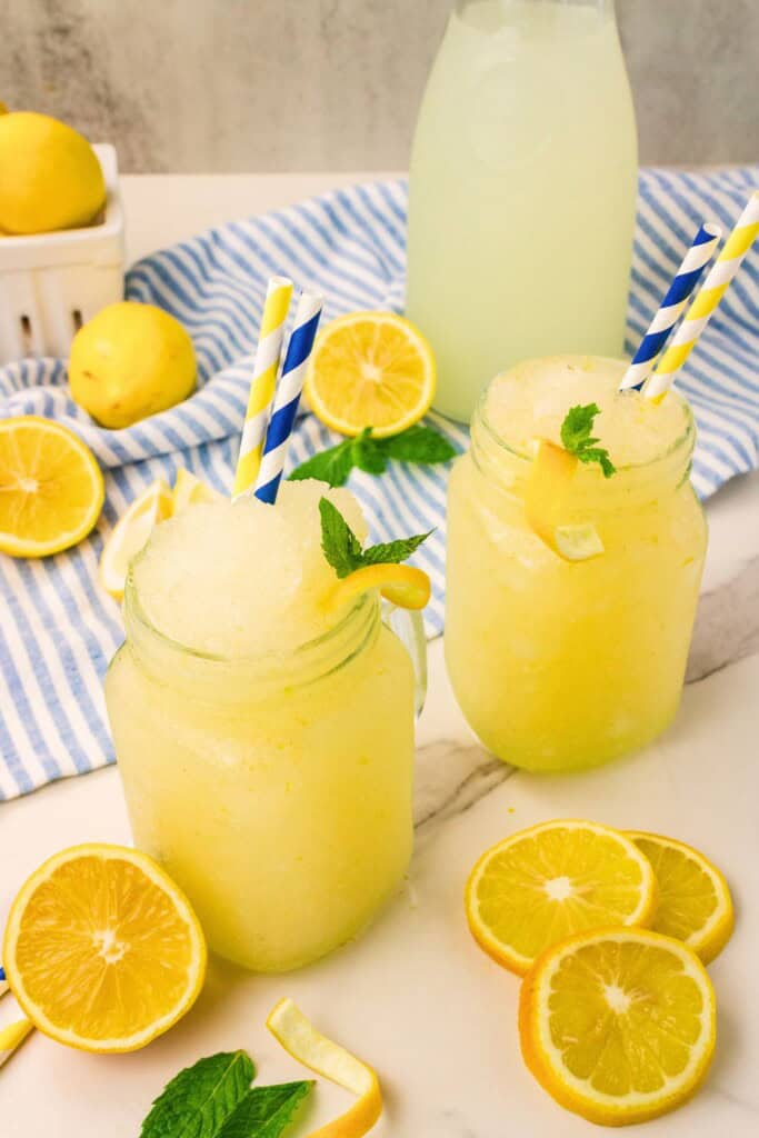 Two lemonade slushies in mason jar mugs with a pitcher of lemonade and fresh lemons