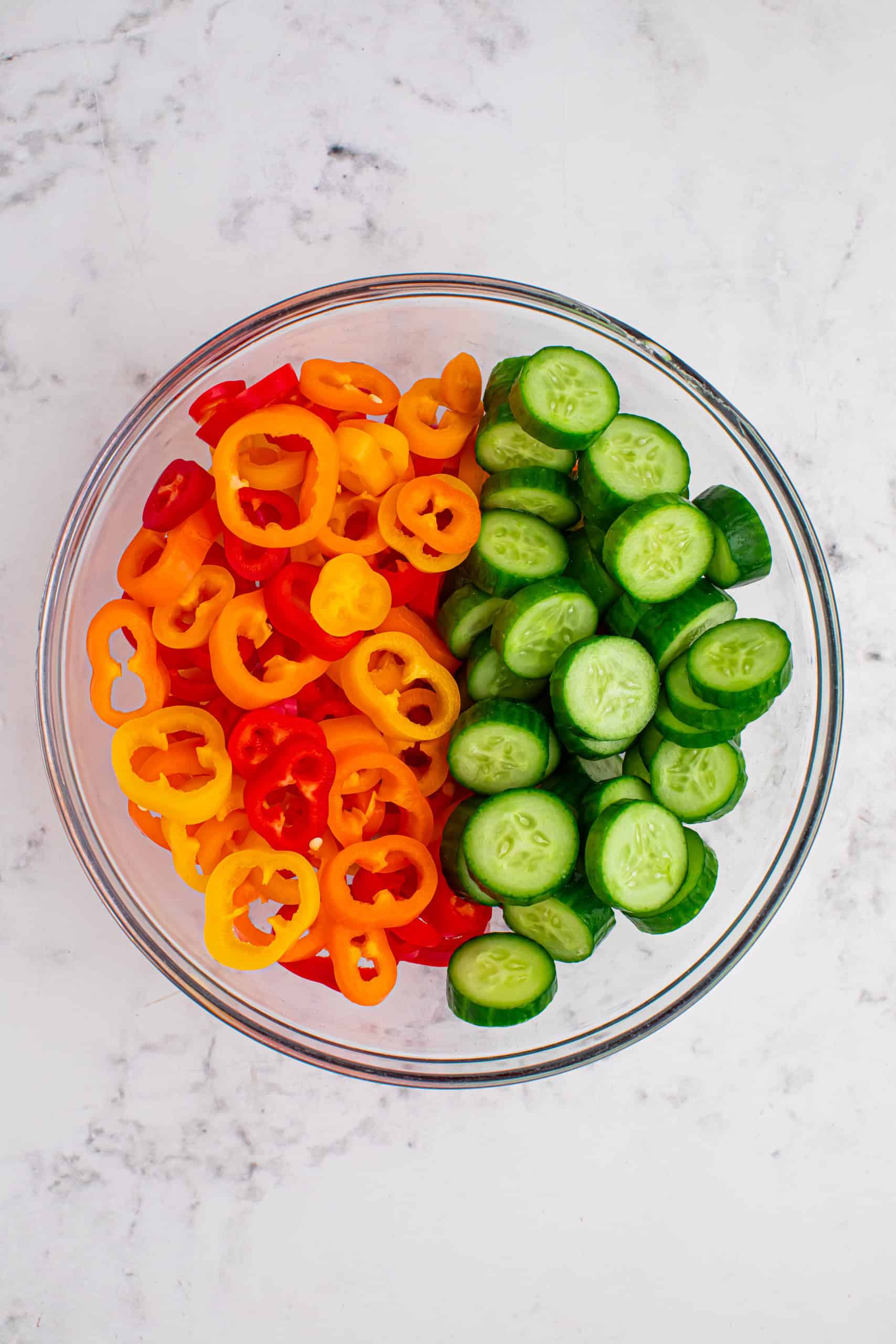 Overhead view of sweet bell pepper rings and sliced mini cucumbers in a glass bowl.