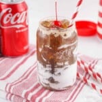 Headon view of fluffy coke in a glass with a cherry on top and a striped straw in it.