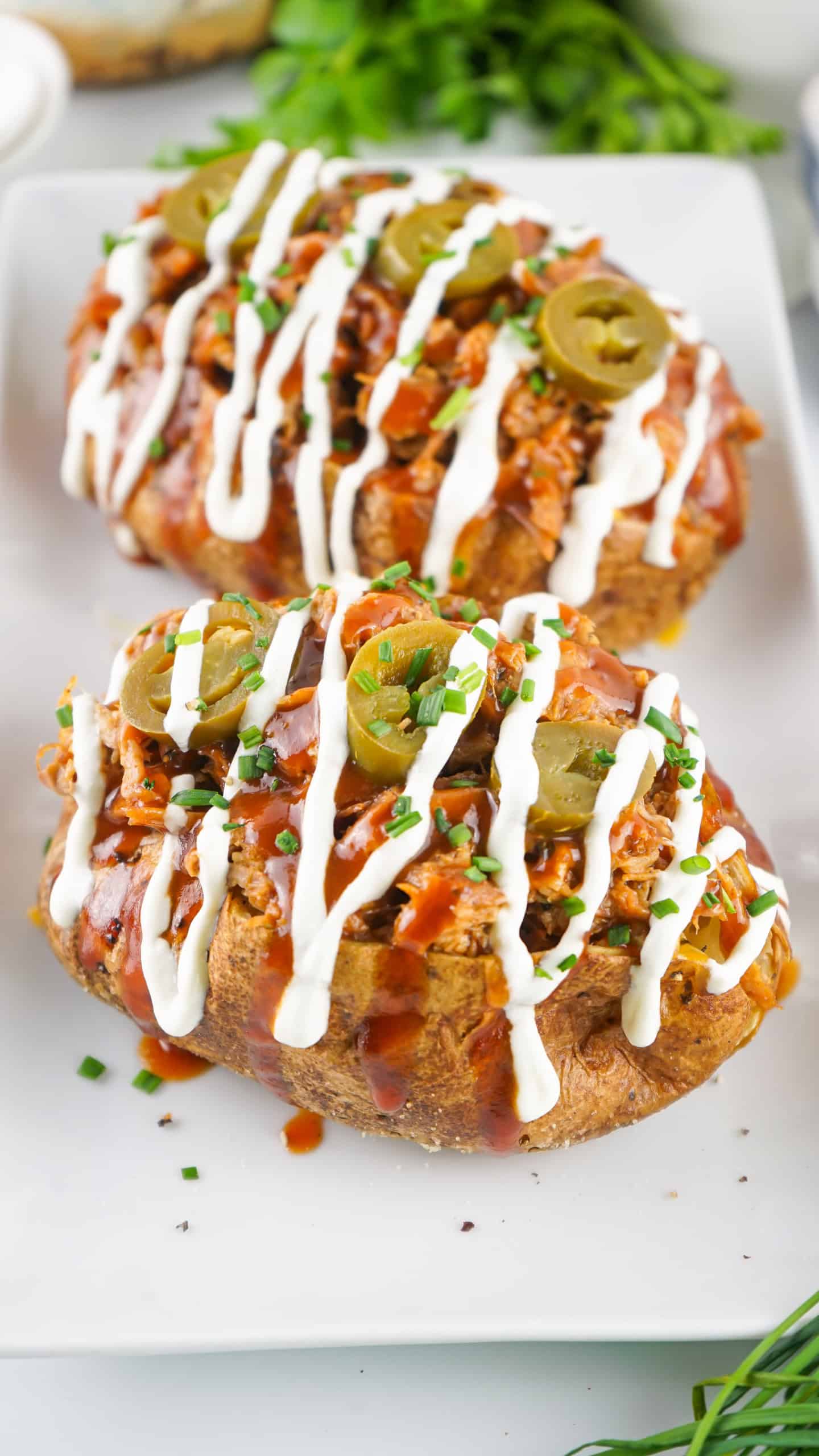 Overhead view of 2 baked potatoes with pulled pork on top, drizzled with BBQ sauce, sour cream and topped with jalapenos.