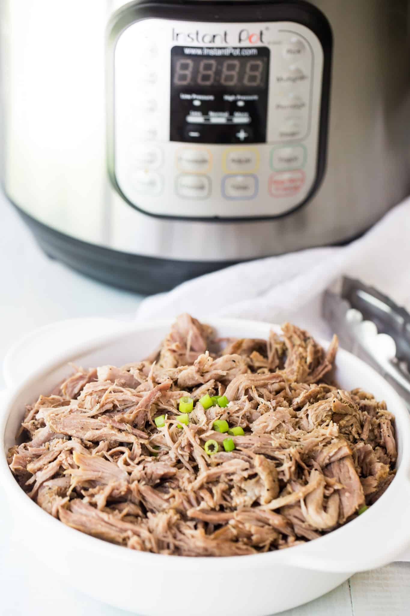 Instant Pot Pulled Pork Butt in white bowl with tongs beside. Photo in front of Instant Pot.