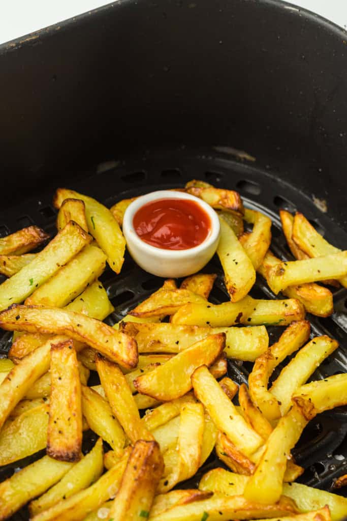 Overhead view of air fryer fries in air fryer basket with small cup of ketchup.