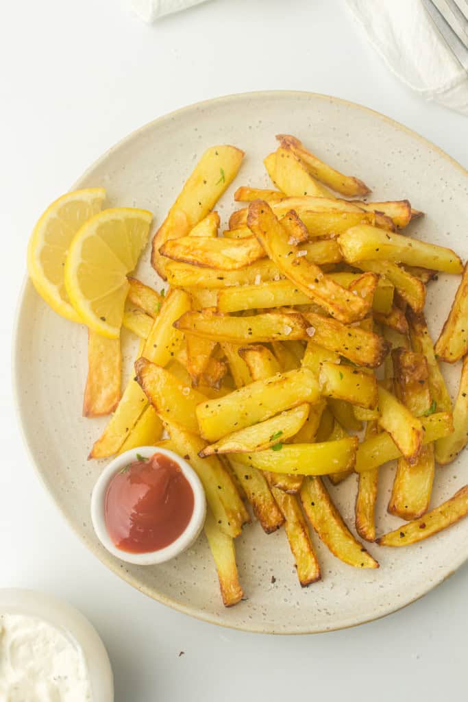Overhead view of homemeade air fryer french fries on a plate with lemon slices and a small bowl of ketchup.