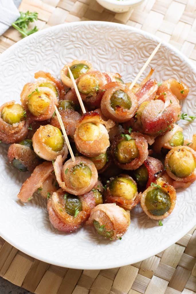 Overhead view of bacon wrapped Brussels sprouts with toothpicks in them in a white bowl.