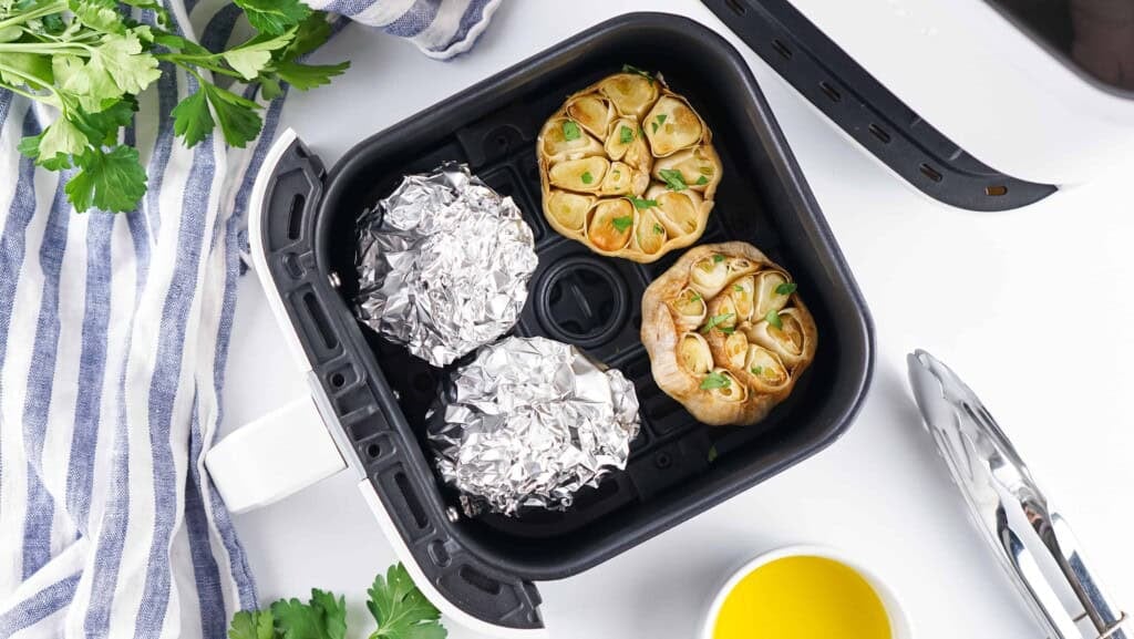 Overhead view of garlic bulbs in air fryer, 2 bulbs wrapped in foil and 2 bulbs with tops cut off.