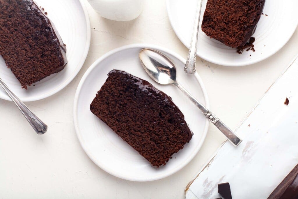 Overhead view of slices of chocolate cake on white plates with silver teaspoons.