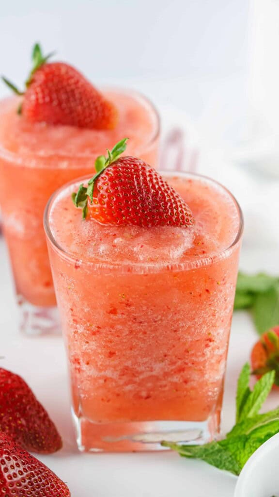 Two tumbler glasses with frozen agua strawberry fresca drinks in them. One glass in front of the otters. Both strawberry Fresca's are topped with fresh strawberries.