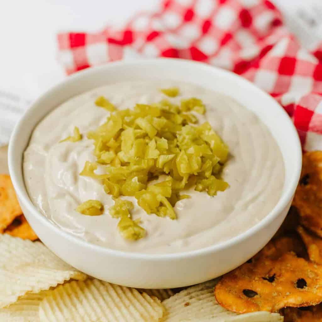 Mississippi Pot Roast Dip in a white bowl with diced up pepperocinis on top and potato chips and pretzels on the side.