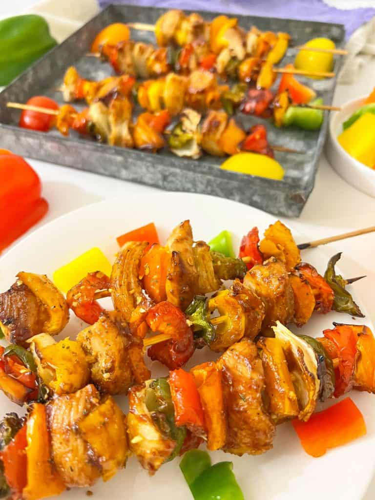 Close-up of chicken and veggie kabobs on a white plate. Tin pan with chicken kabobs on it in the background.