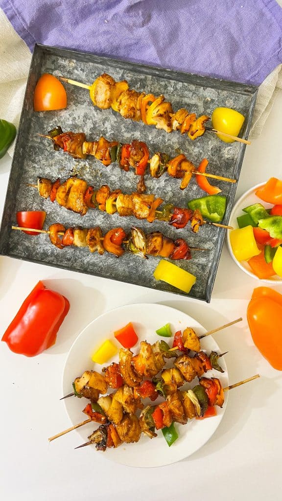 Overhead view of BBQ chicken kabobs. 3 on a white plate and 4 on a tin pan beside the plate.
