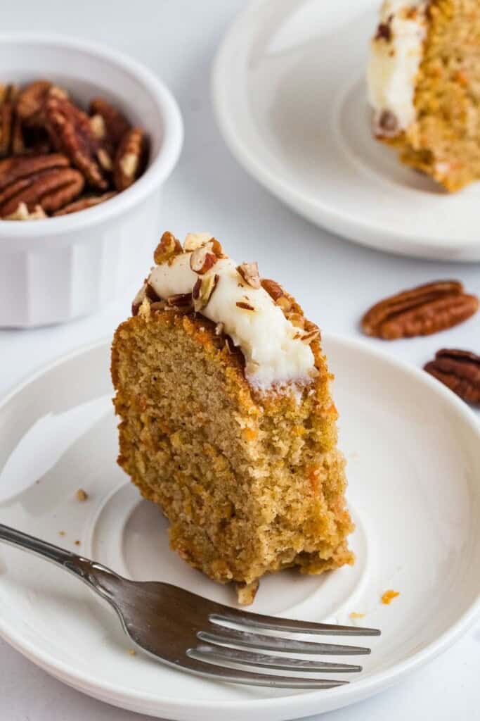 Slice of copycat Nothing Bundt carrot cake on a white plate with a fork and bowl of pecans
