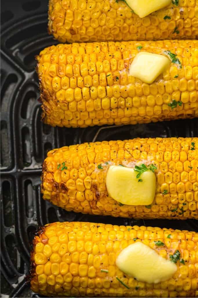 Air fryer corn on the cob in air fryer basket with melted butter, flaky salt, and a sprinkle of fresh parsley.